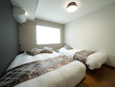 Comfortable twin bedroom at Canal Villa Otaru with white bedding, geometric patterned runners, natural lighting, and modern ceiling fixture.