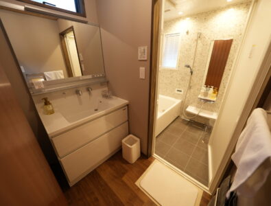 Modern bathroom at Enishiya Izumo Villa with double vanity, glass-enclosed shower, soaking tub, and premium amenities in warm lighting.