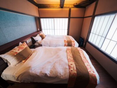 alt="Traditional Japanese bedroom at Enishiya Izumo Villa with twin beds, exposed wooden beams, shoji screens, and elegant floral bedding accents.