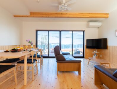 Bright open-plan living space at Ocean Villa Naruto with natural wood furnishings, ocean view terrace, dining table, and modern leather seating.